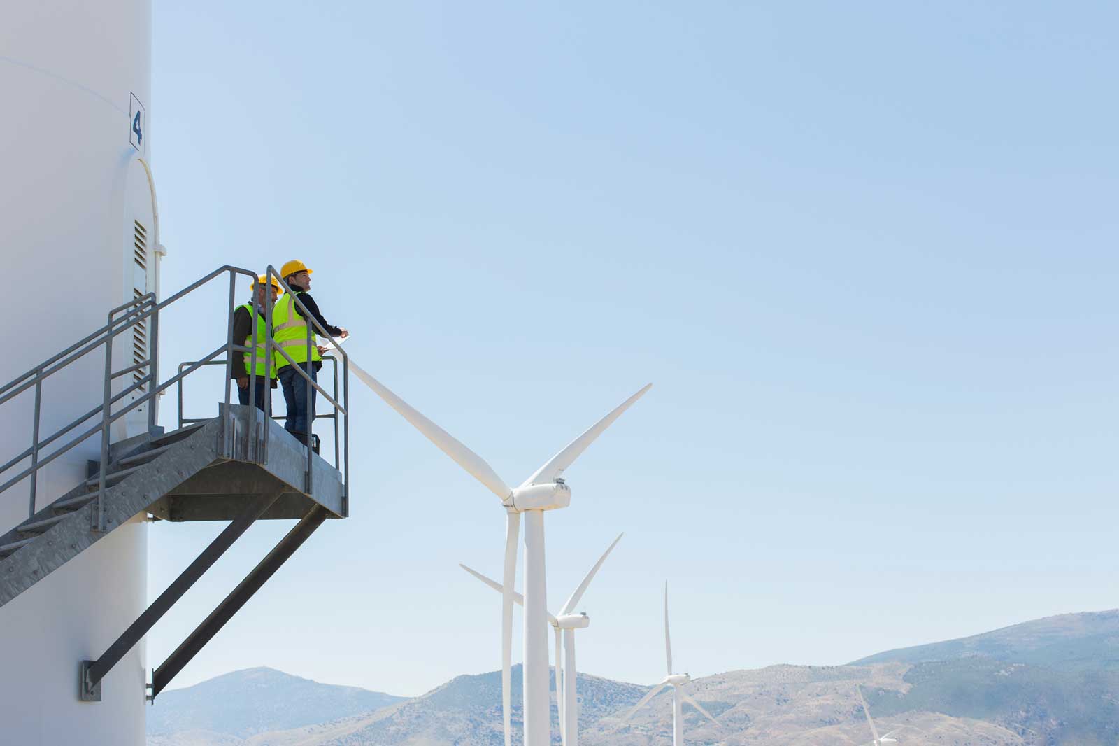 Two workers in safety gear stand on a platform beside a wind turbine, overlooking a landscape of additional turbines and mountains.