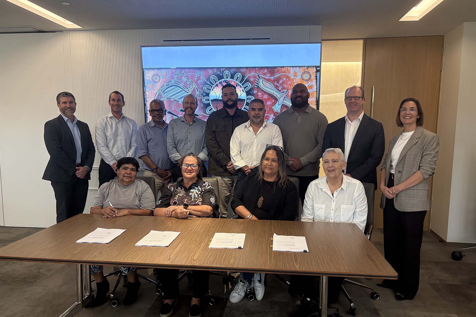 Thirteen professionally dressed adults pose around a wood conference table with papers, smiling in a modern meeting room with a colorful mural behind them.