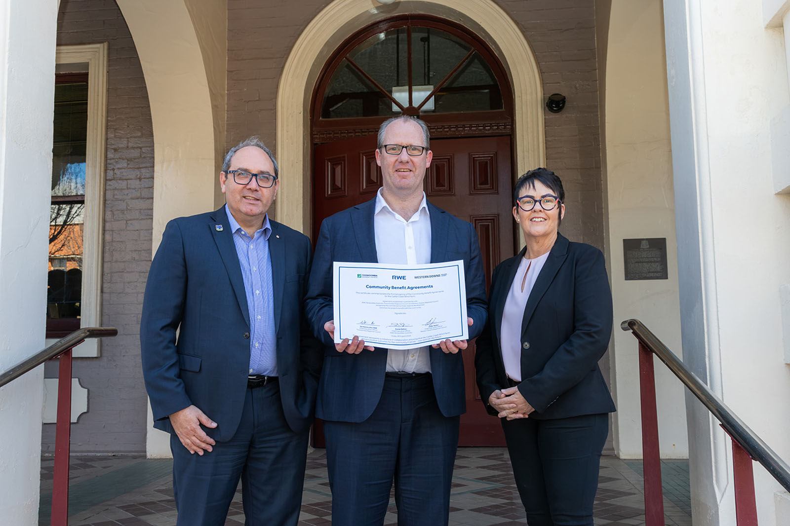 Three people wearing suits stand on tiled steps in front of an arched red door, the central man holding a certificate while both companions smile.
