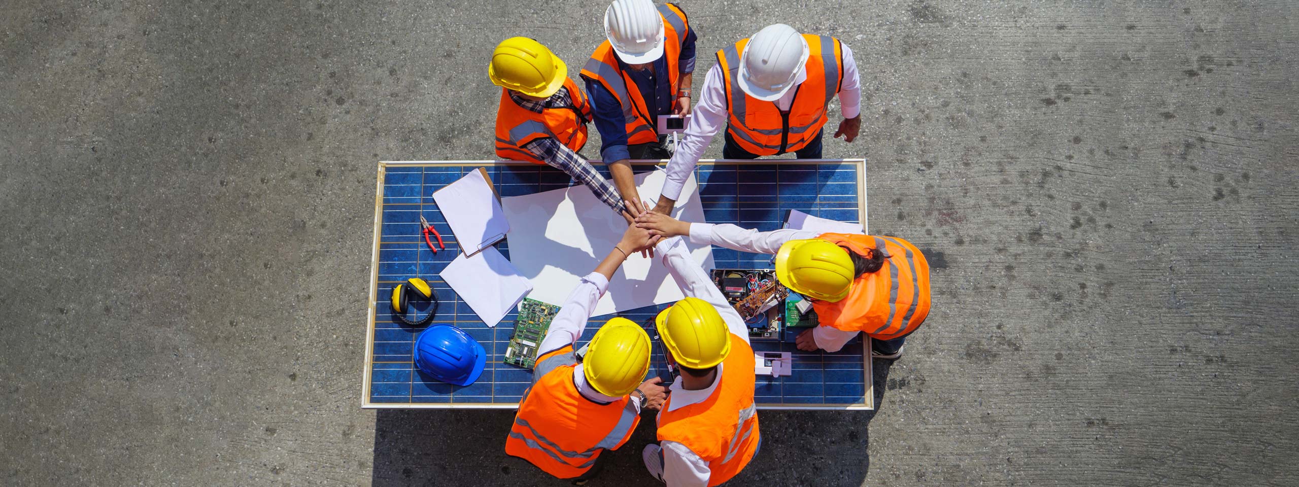A group of construction workers in hard hats and safety vests collaborate at a table covered with papers and tools.