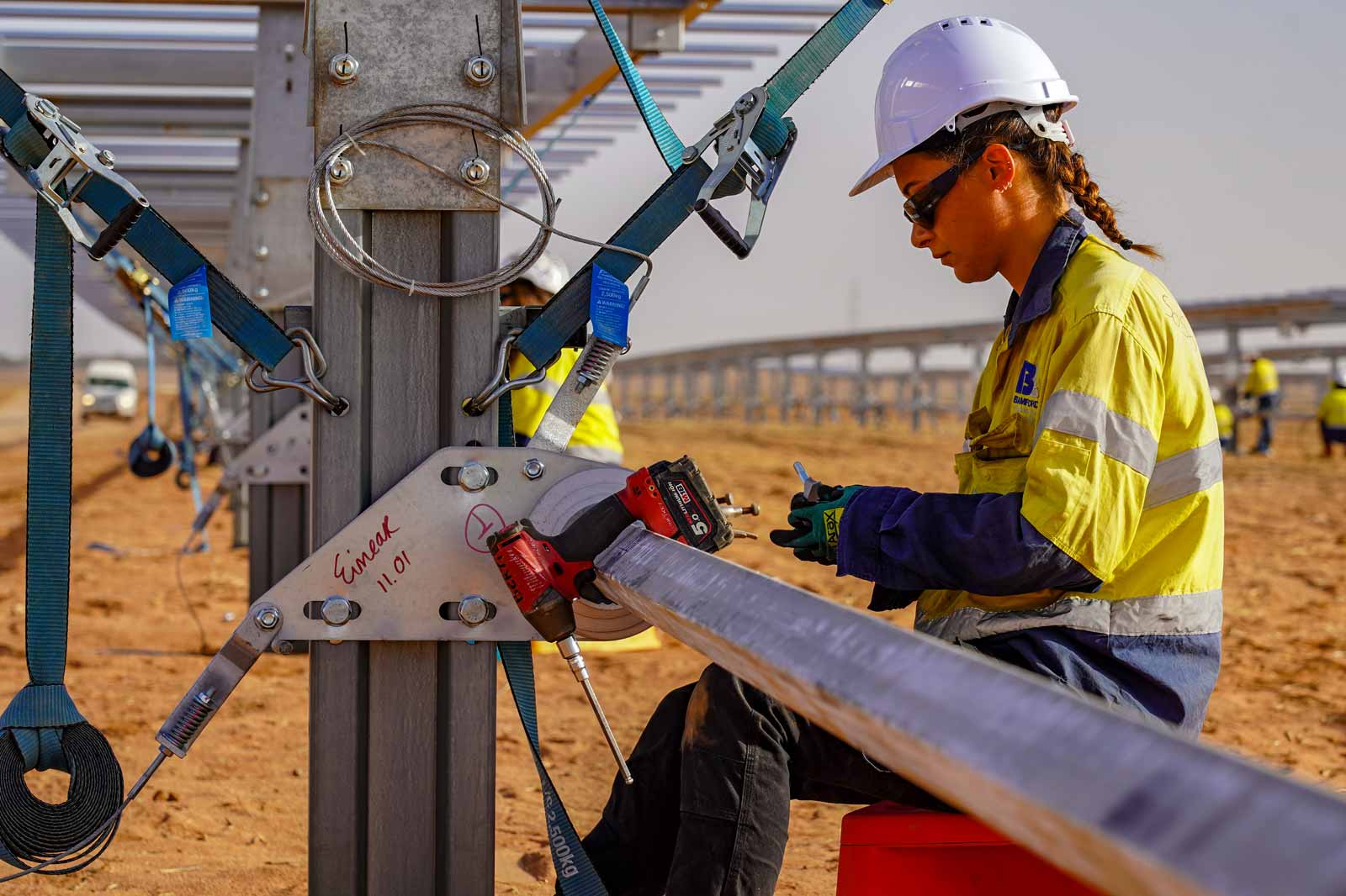 A worker in safety gear uses a tool on a metal structure at a solar panel installation site in a desert landscape.