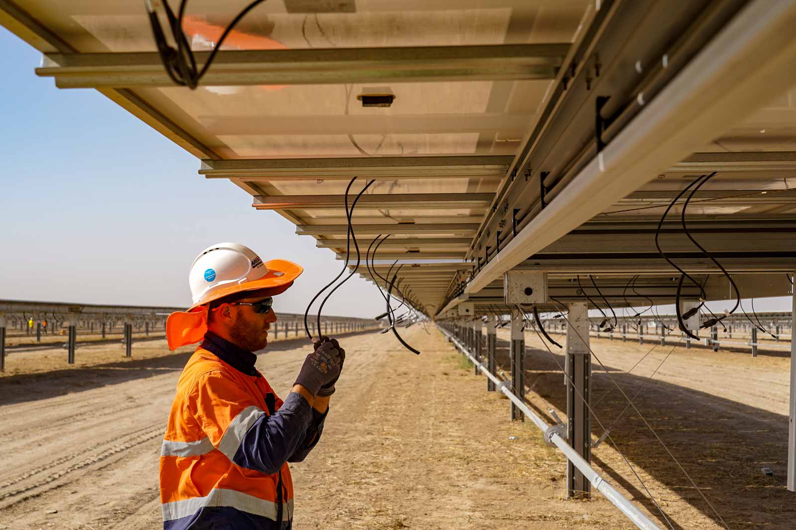 A worker in an orange and blue uniform inspects solar panels, with cables visible above on a sunny day in a solar field.