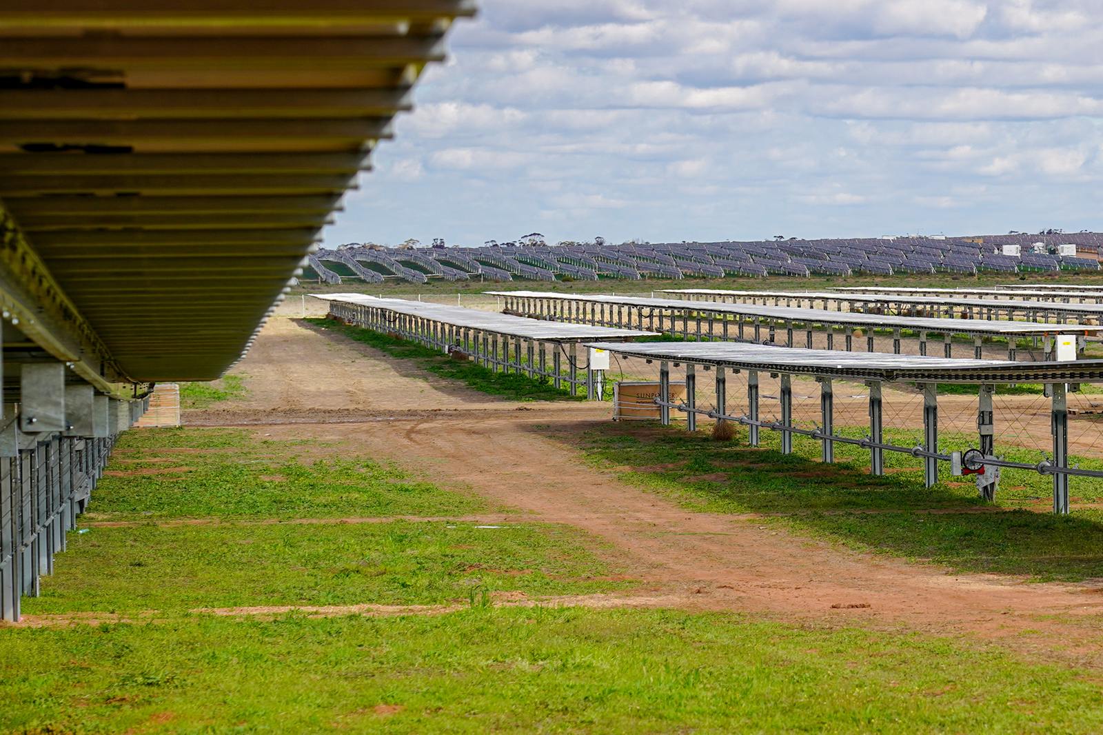 A solar farm with rows of solar panels under a cloudy sky, green grass and dirt paths visible in the foreground.