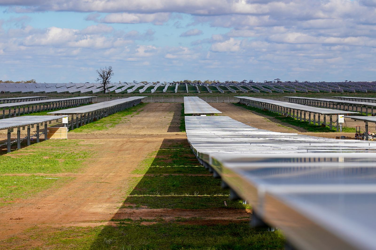 A wide view of a solar farm with rows of solar panels under a cloudy sky. Green grass and dirt path visible.