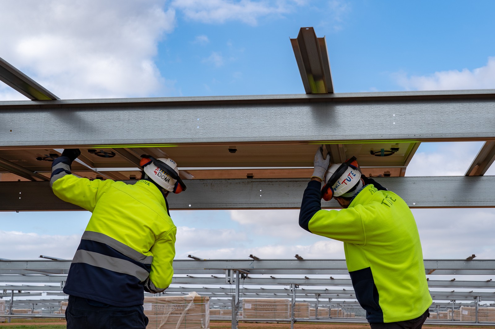 Two workers in high-visibility jackets install solar panels under a blue sky with scattered clouds.