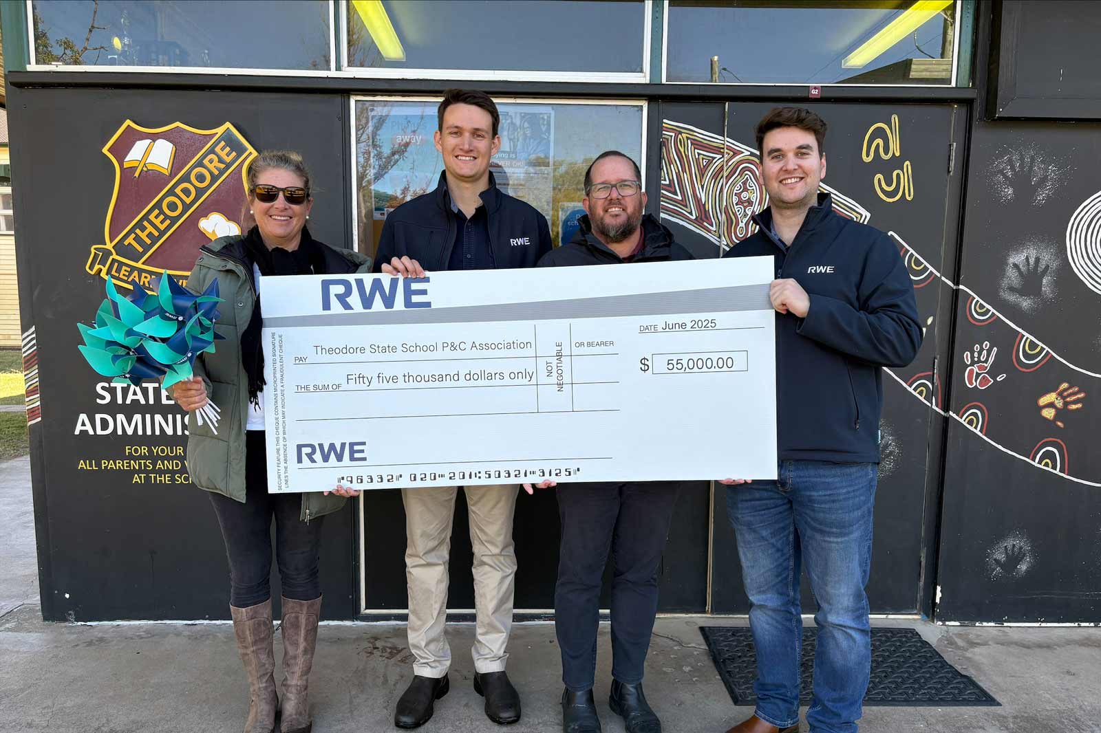 A group of people hold a large cheque for fifty-five thousand dollars in front of a school with colourful murals.