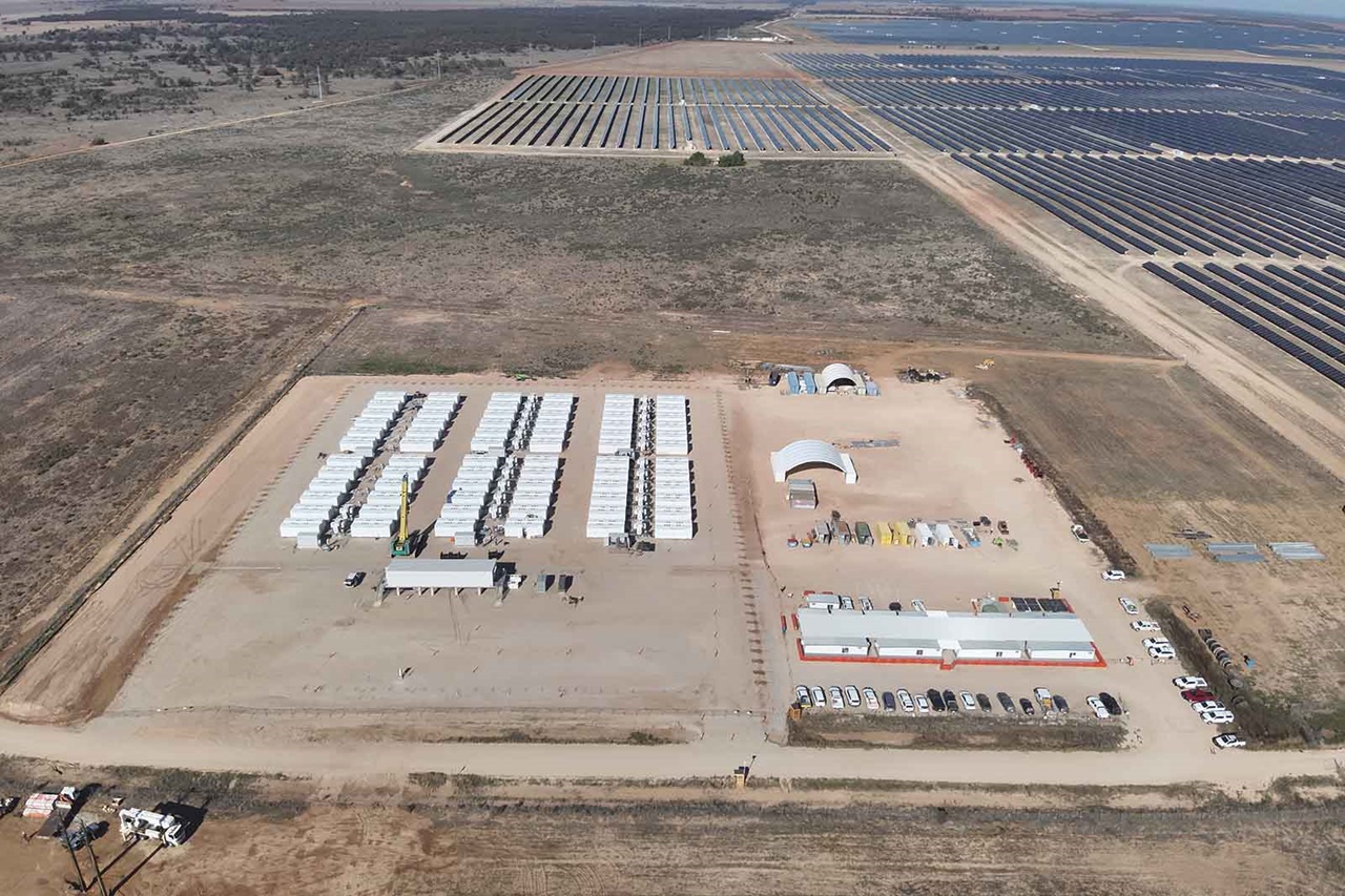 Aerial view of a dusty rectangular work compound with white modular units, parked trucks by beige structures, and distant dark solar panel arrays.