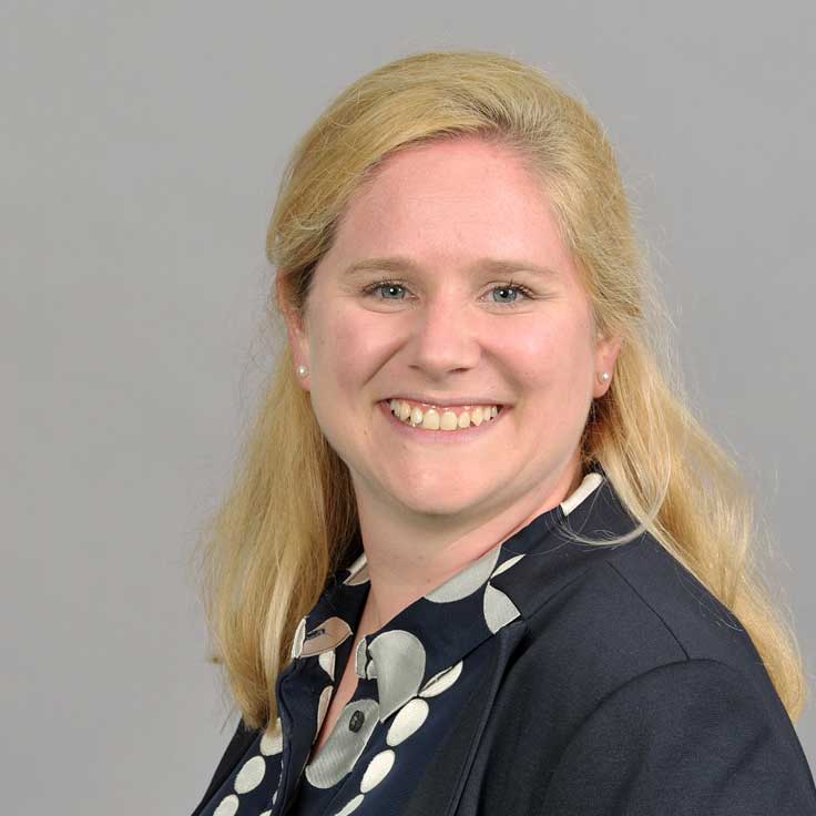 Woman with blonde hair and earrings, wearing a dark jacket with a spotted pattern. Portrait of Sarah Knauber.