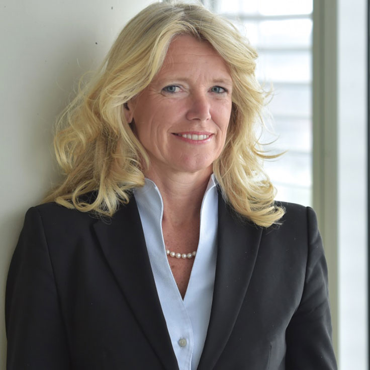 A woman with blonde, curly hair wearing a black blazer and a pearl necklace stands in front of a window. Portrait of Stephanie Schunck.