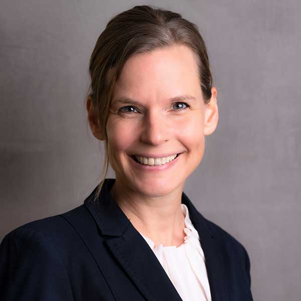 A woman is wearing a navy blazer over a white blouse, standing against a neutral grey background.
