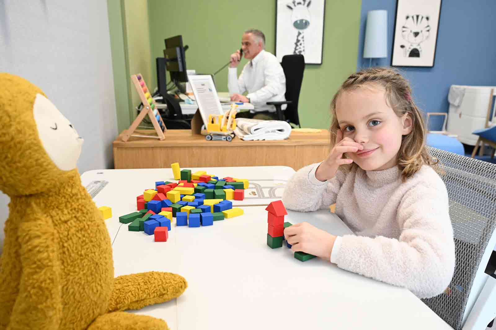 A child sits at a table with colourful building blocks, while a plush monkey is nearby. In the background, an adult is working.