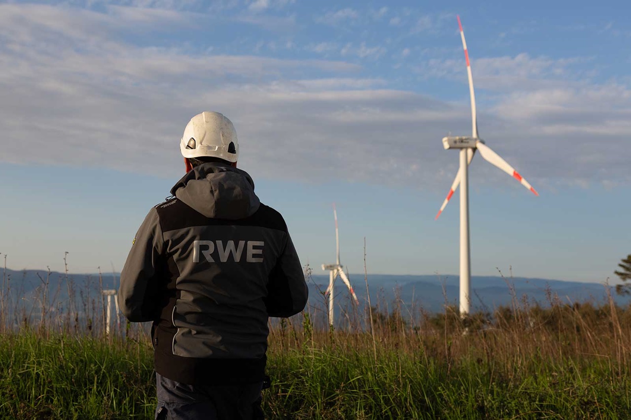 A worker in a safety helmet and jacket marked RWE stands on a grassy hilltop facing wind turbines under a blue sky with distant hills.